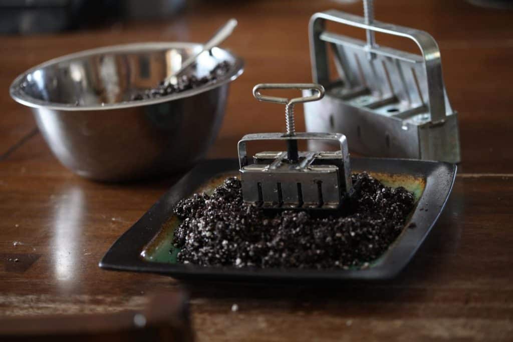 square plate with soil, two metal soil block makers, and a steel bowl full of soil with a spoon