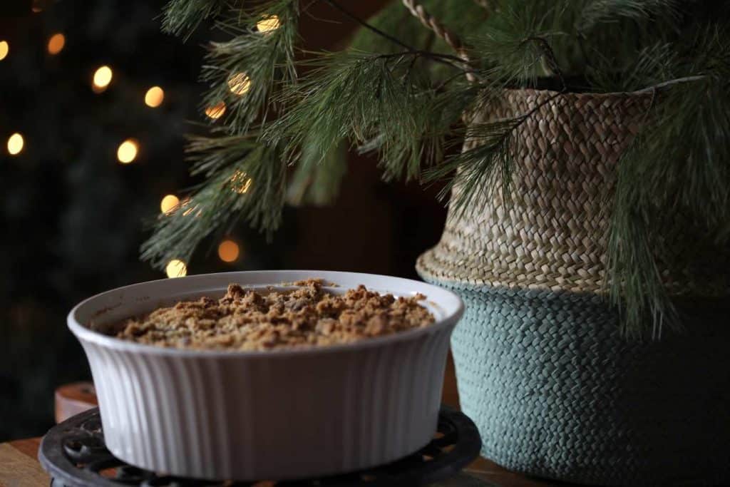 white casserole dish on a trivet, with sweet potato casserole, next to a basket with pine boughs and twinkling lights in the background, showing how to make a sweet potato casserole
