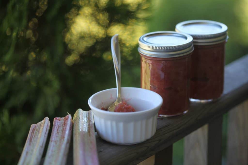 two mason jars with rhubarb jam on a wooden railing next to a white bowl with jam and a silver spoon, as well as several pieces of chopped rhubarb