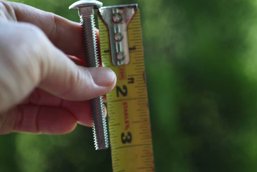 a hand holding a three inch bolt next to a measuring tape