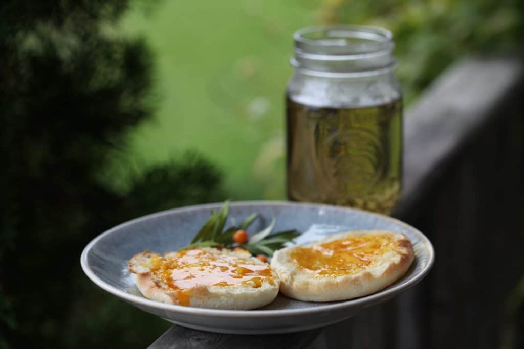 sea buckthorn jam on an English muffin on a plate, with sea buckthorn tea in a mason jar on a wooden railing