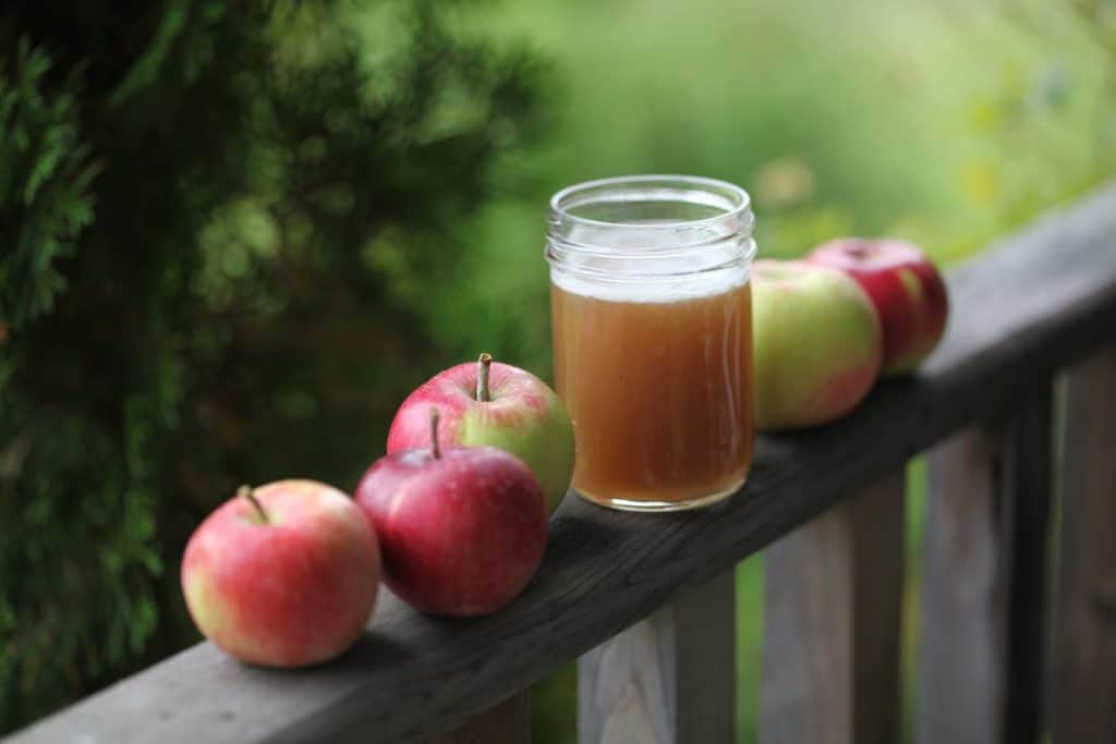 a glass of freshly squeezed apple cider on a wooden railing with five red  and yellow apples