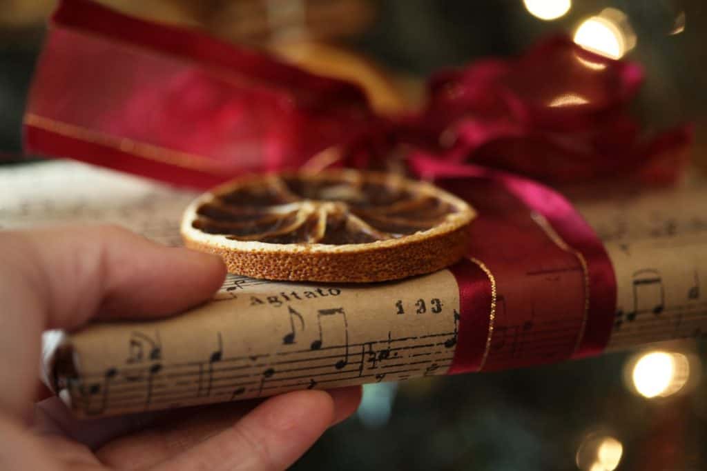 a hand holding a gift wrapped in music note paper, decorated with a dried orange slice and a red ribbon