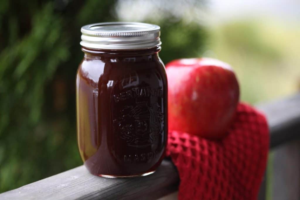 easy to make apple cider syrup recipe, with a mason jar full of dark brown syrup next to a red apple on a red cloth, on a grey wooden railing