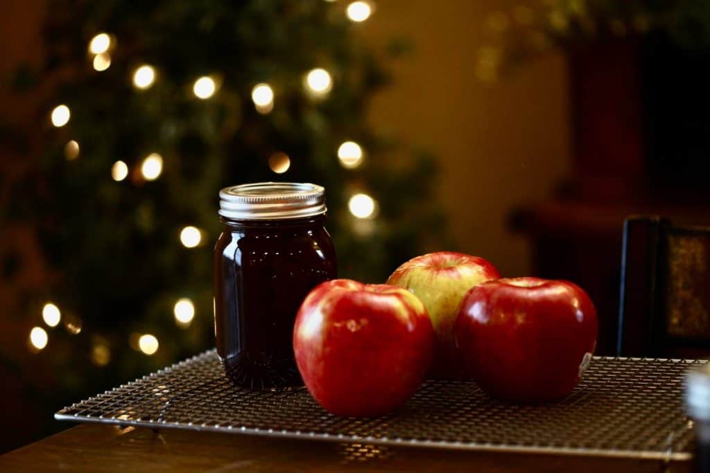 freshly made apple cider syrup in a mason jar on a cooling rack next to three red apples with white lights in the background