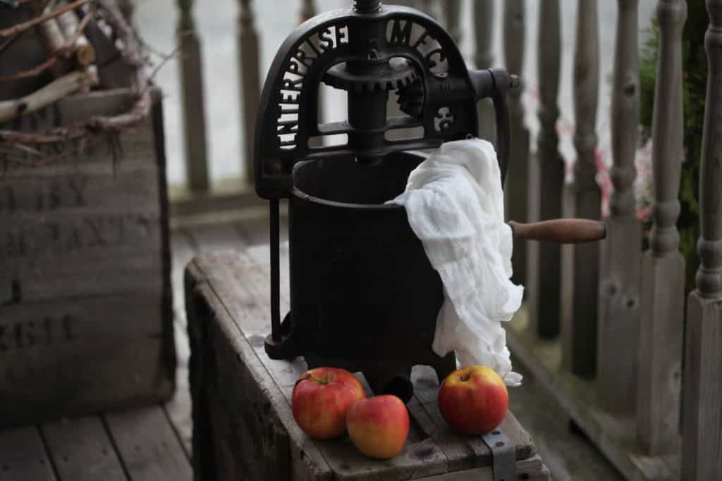 an old apple press on a wooden crate with cheesecloth hanging out, next to three red and yellow apples