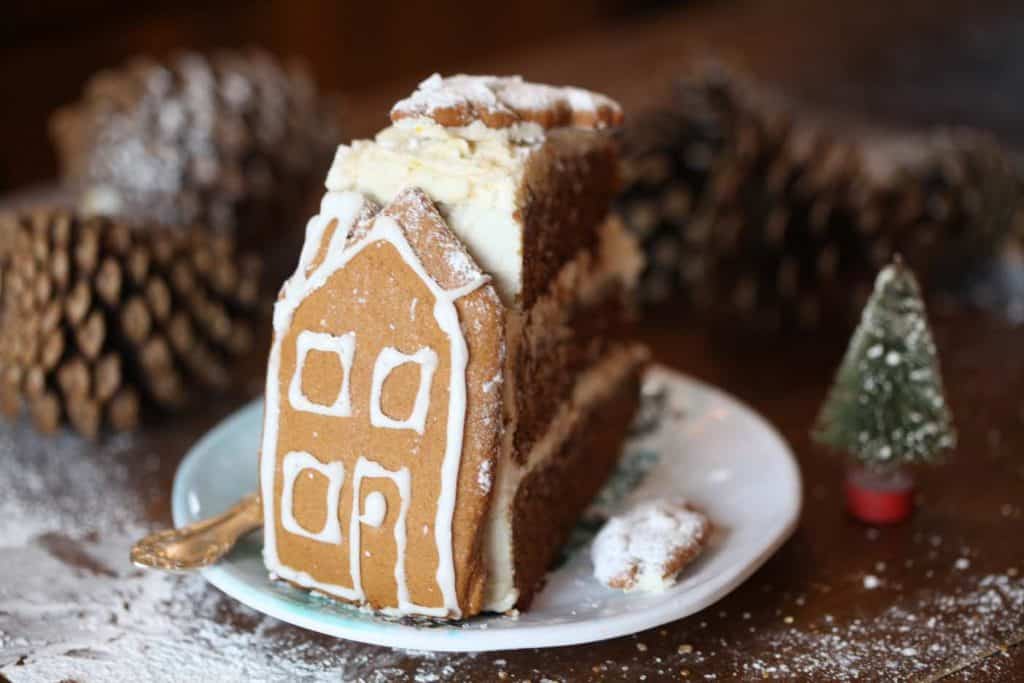 a slice of gingerbread cake with cream cheese frosting on a small plate surrounded by brown pine cones and decorated with a gingerbread house cookie