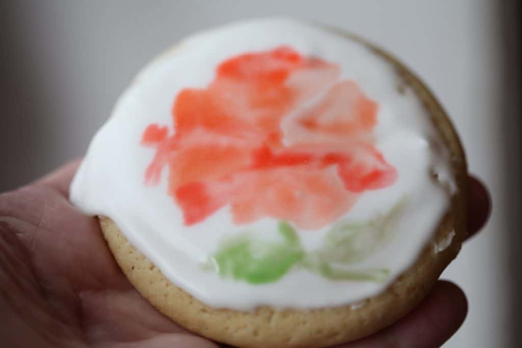 a hand holding a cookie showing a flower painted with edible watercolor on a sugar cookie
