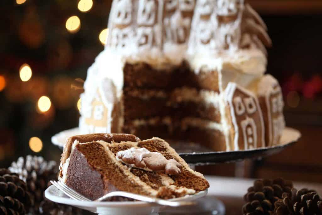 gingerbread cake with cream cheese frosting, decorated with gingerbread house cookies, with a slice of cake cut out and sitting on a plate in front of the cake
