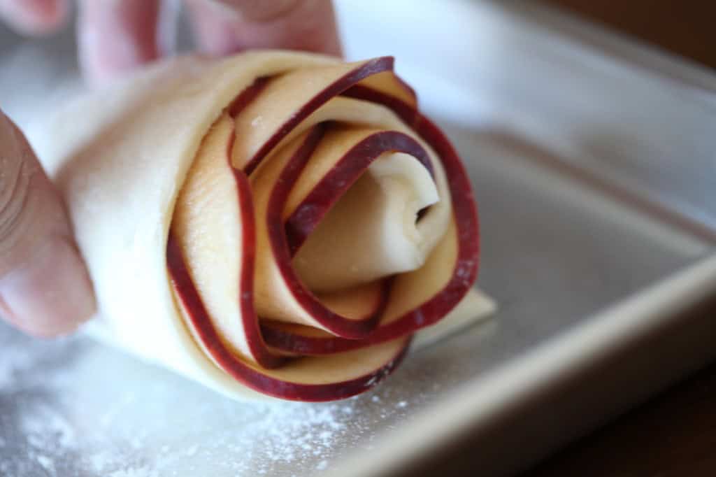 Close-up of a hand shaping a pastry into a rose using thin slices of red apple layered in rolled dough on a floured baking sheet.