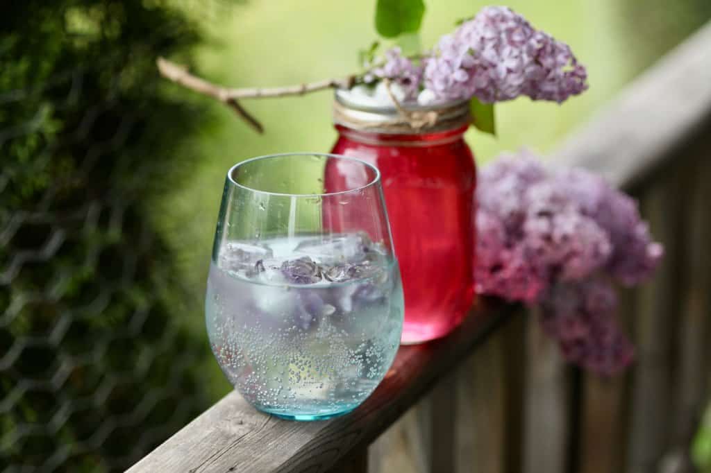 a glass with a carbonated beverage in front of a mason jar filled with red coloured syrup and topped with purple lilacs, on a wooden railing