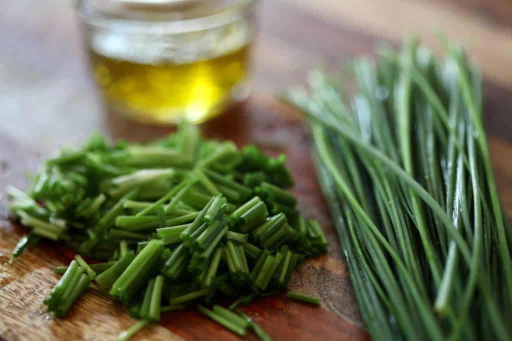 chopped chives next to a bundle of chives and a glass of olive oil on a wooden cutting board