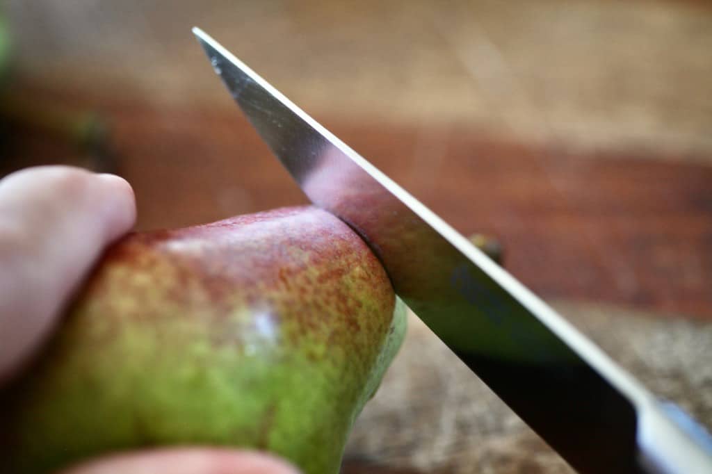 a hand holding a pear and cutting off a stem with a paring knife