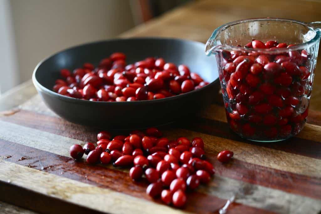 rose hips in a black bowl, a clear glass measuring cup, and on a wooden cutting board, in preparation to make a rose hip syrup recipe