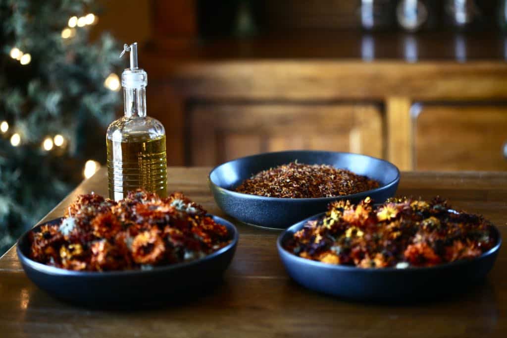 three bowls of dried calendula flowers and a bottle of olive oil on a wooden table