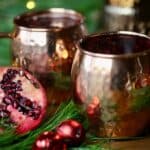 Christmas Moscow mules in copper mugs next to a halved pomegranate, fir boughs, and red Christmas ornaments