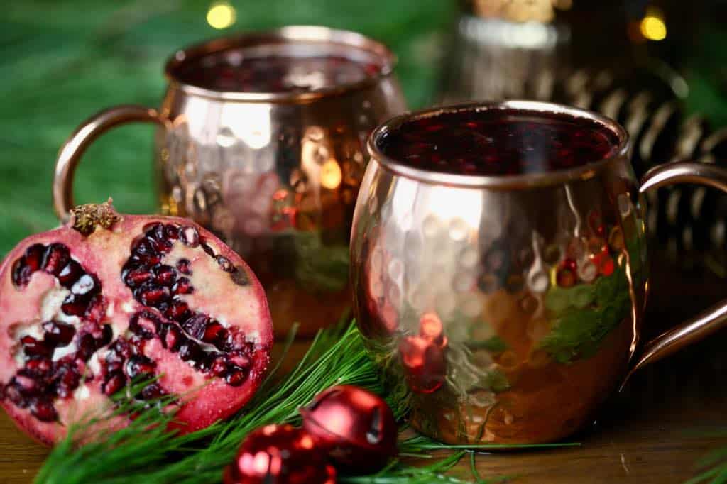 Christmas Moscow mules in copper mugs next to a halved pomegranate, fir boughs, and red Christmas ornaments