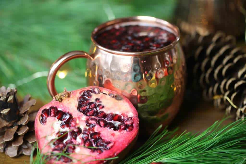 a copper mug filled with Christmas mule, next to pine boughs and pine cones, and a cut pomegranate half