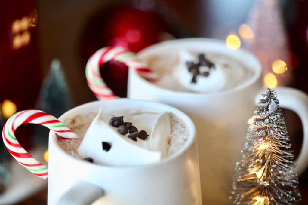 two white mugs with hot chocolate topped with whipped cream and chocolate chips, with a candy cane stir stick