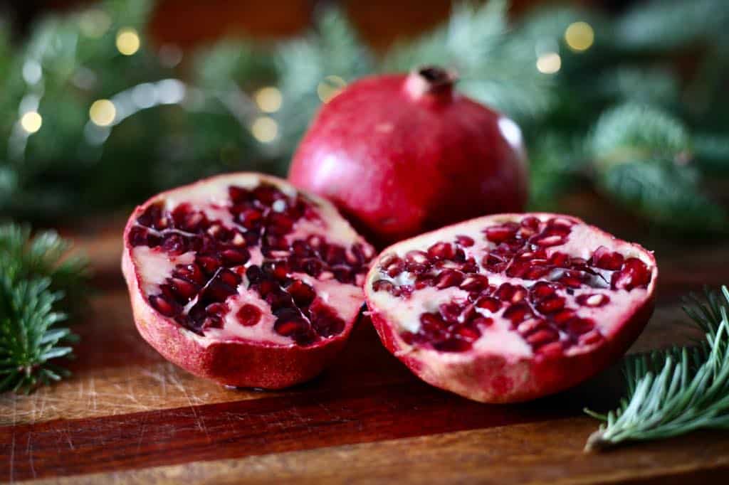 pomegranates on a wooden cutting board in front of green boughs, with one pomegranate cut in half